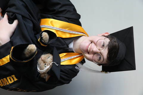 GH Jessie Meiers poses in their graduation robes with their orange cat in their arms; the cat is also dressed in graduation robes.