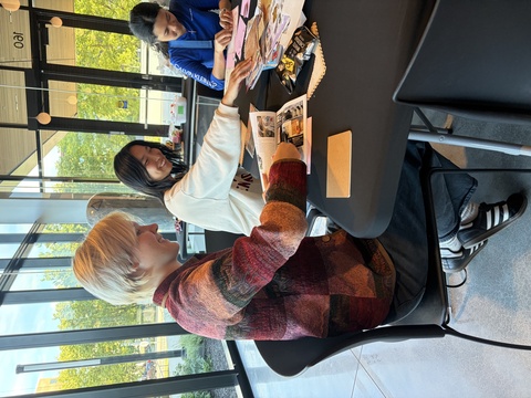Three students sit around a table, actively collaging with piles of materials.