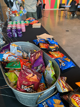 A close up photo of the table of snacks and drinks at the Art of Voting event, featuring colorful single serve bags of chips and sparkling water.