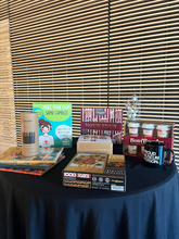 cocktail table with a black cloth over it. On top of the table are an assortment of bingo prizes consisting of colored pencils and coloring books, hot chocolate and mug set, puzzle, coaster making set, and a popcorn kit 