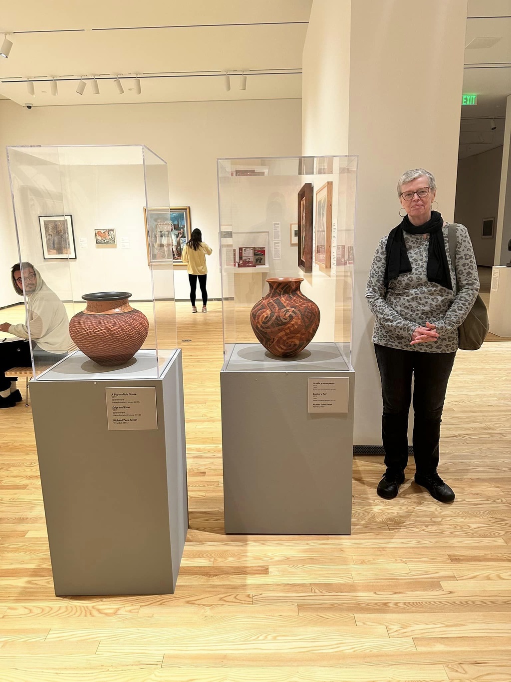 A woman with short grey hair wearing black pants and a grey, long-sleeve top with a black scarf stands next to two red, ceramic pots