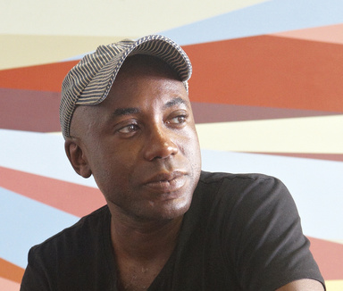 A Black man in a dark t-shirt and beige ball cap in front of a mural painted with geometric shapes in bright colors.