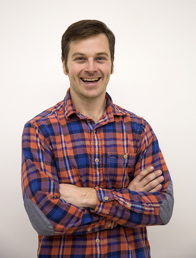 Smiling man with short, dark hair and wearing a red and blue plaid shirt. His arms are folded across his chest.
