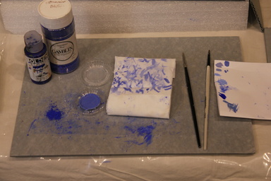 Art conservator work surface with bottles of pigment, loose pigment, paint brushes, and blotting paper with deep blue marks.