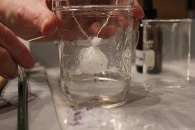 A small cheesecloth sachet suspended in a glass jar of solvent with string