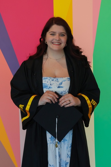 A photo of Alexis Belme in her graduation cap and gown; she poses, smiling, in front of the multi-colored mural in the Stanley Museum of Art lobby.