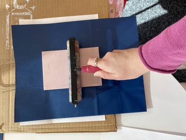 A photo of a print being made: Annelies is rolling a brayer over the back of the linoleum block, which is face down on the blue paper bag, transferring the image in white ink onto the bag.