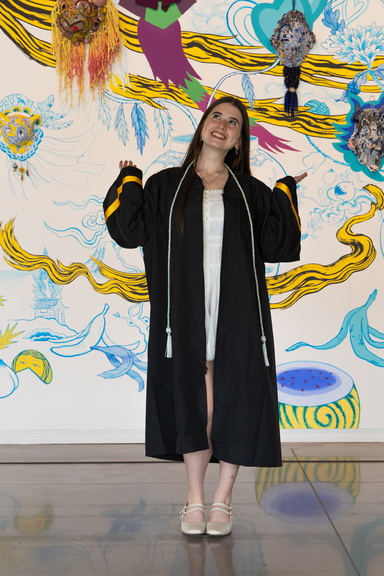 Gallery Host and Campus Engagement Coordinator Annelies Knight poses in her graduation gown in front of Jiha Moon's mural.