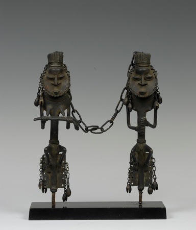 Two angular metal figurines with detailed faces and headpieces are connected by a chain, with additional chains hanging from their heads and waists. The figure on the left represents a female and the one on the right a male. They are affixed onto a dark rectangular base, placed in front of a plain grey backdrop.