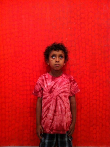 A young boy with dark curly hair, wearing a bright red tie-dye pattern shirt, stands in front of Steve Erickson's monumental red painting, "Earth," and glances upward.
