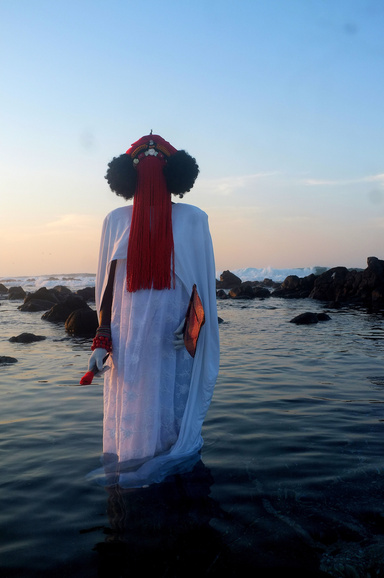 Photograph of a figure in a white robe and elaborate red beaded headdress that covers the face, standing ankle-deep in the ocean, holding objects in each hand. In the background is a pale sky during sunset with calm water and rocks along the shoreline. 