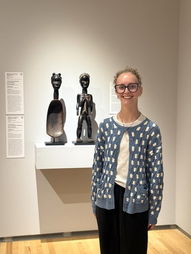 A woman with curly blonde hair and glasses, wearing a blue cardigan with a white dot pattern, stands smiling in a museum gallery beside a wooden feast spoon carved with a woman's head and a dark wooden figure, displayed on a pedestal with wall labels for the Dis/Comfort exhibition.