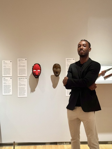 A man in a black blazer and tan pants stands with arms crossed in a museum gallery, beside two African masks mounted on the wall, one with a red face and animal fur trim, the other a smaller dark wooden mask, with wall labels and a display case visible nearby.