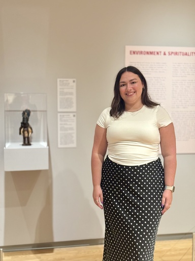 A woman with dark hair, wearing a cream top and a black-and-white polka-dot skirt, stands smiling in a museum gallery near a display case holding a small carved figure, beneath a red 'Environment and Spirituality' section header.