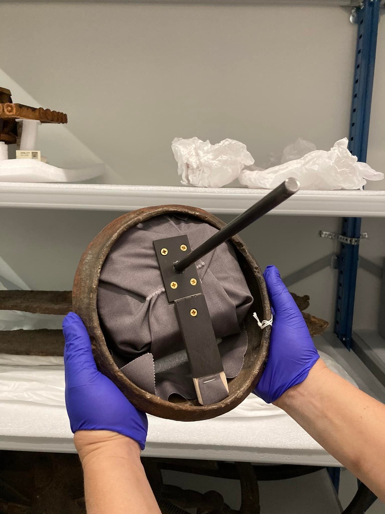 Two hands wearing purple gloves hold a wooden face mask. Inside the back of the mask is a padded form connected to a metal bracket. There is a metal rod protruding from the bracket that will connect with a wall-mounted bracket in the gallery.