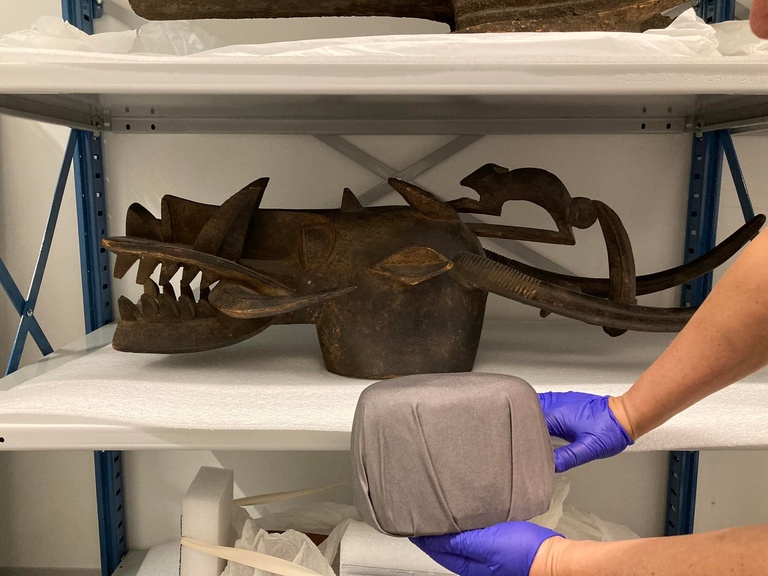 A wooden mask shaped like a dragon (fire-breather) sits on a shelf. In front, two hands in purple gloves hold a padded form wrapped in grey cloth that will be used as an insert to stabilize the mask on its wall mount.
