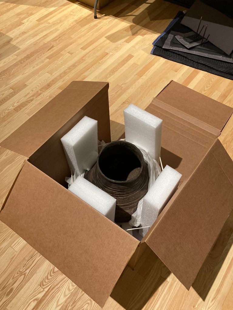 Large ceramic pot in a cardboard packing box is surrounded and protected by white foam packing material.