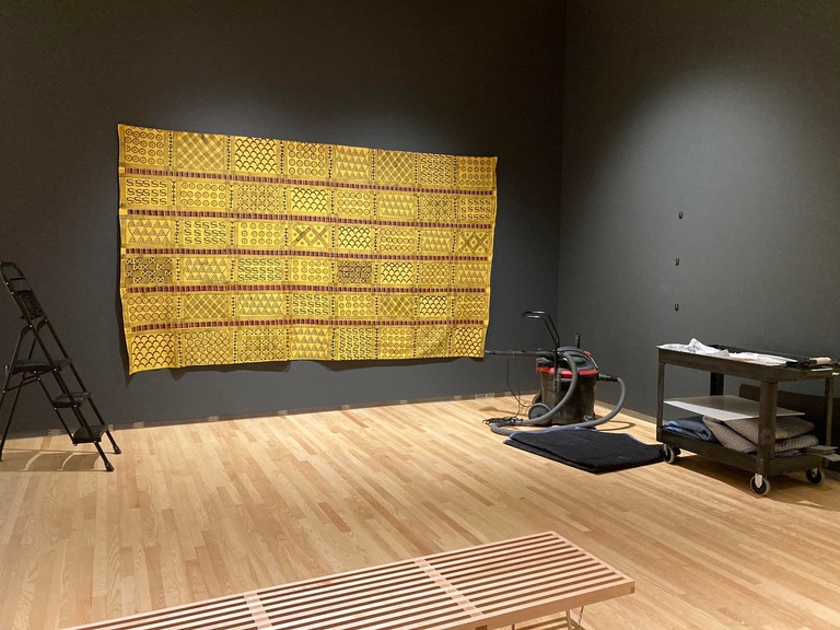 Yellow kente cloth hangs on a dark grey wall with a vacuum cleaner and wheeled cart in the foreground.