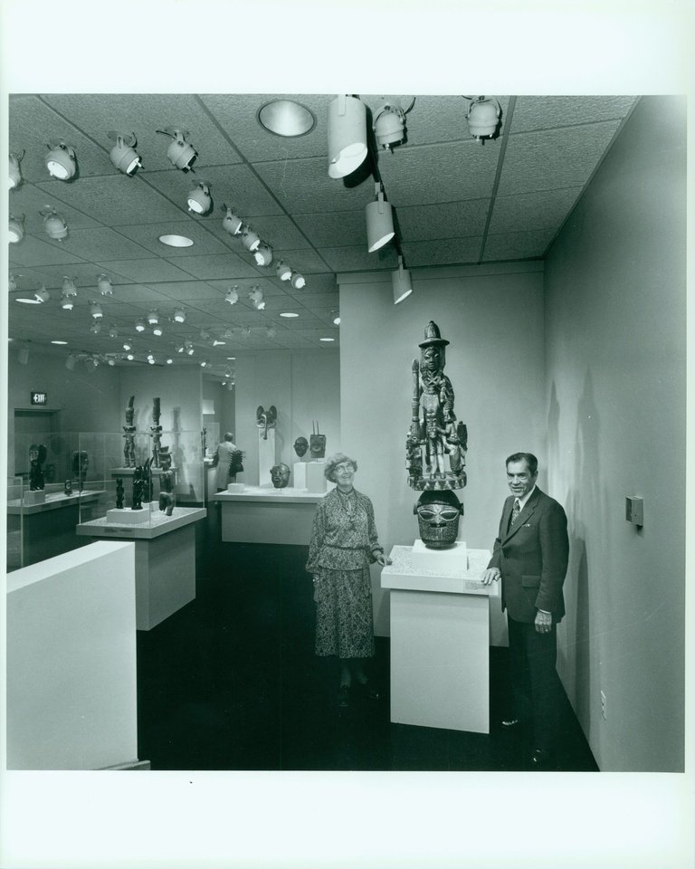 A woman and man stand in a museum gallery on either side of an African (Epa) wooden mask. There are other African art objects in the background.