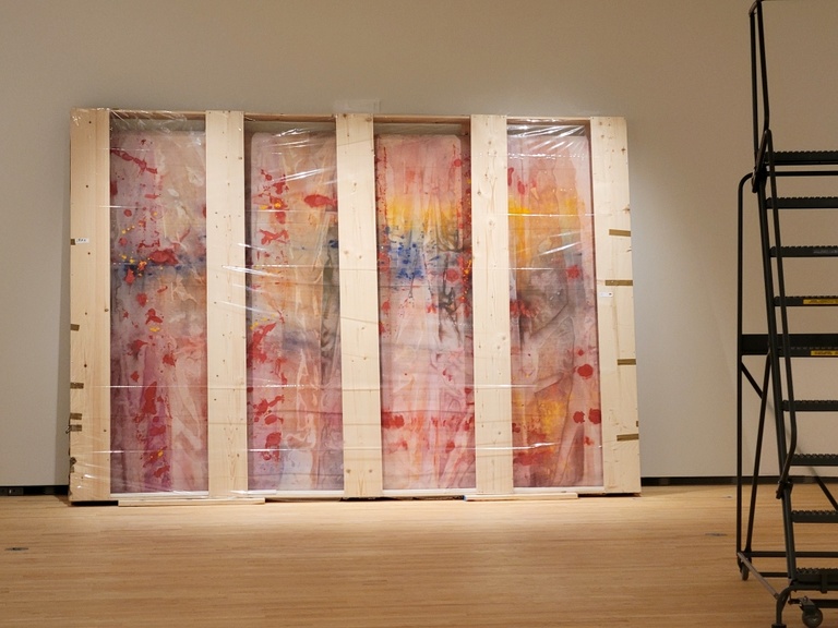 Large, multi-colored painting in it's wooden crate leans against a gallery wall.