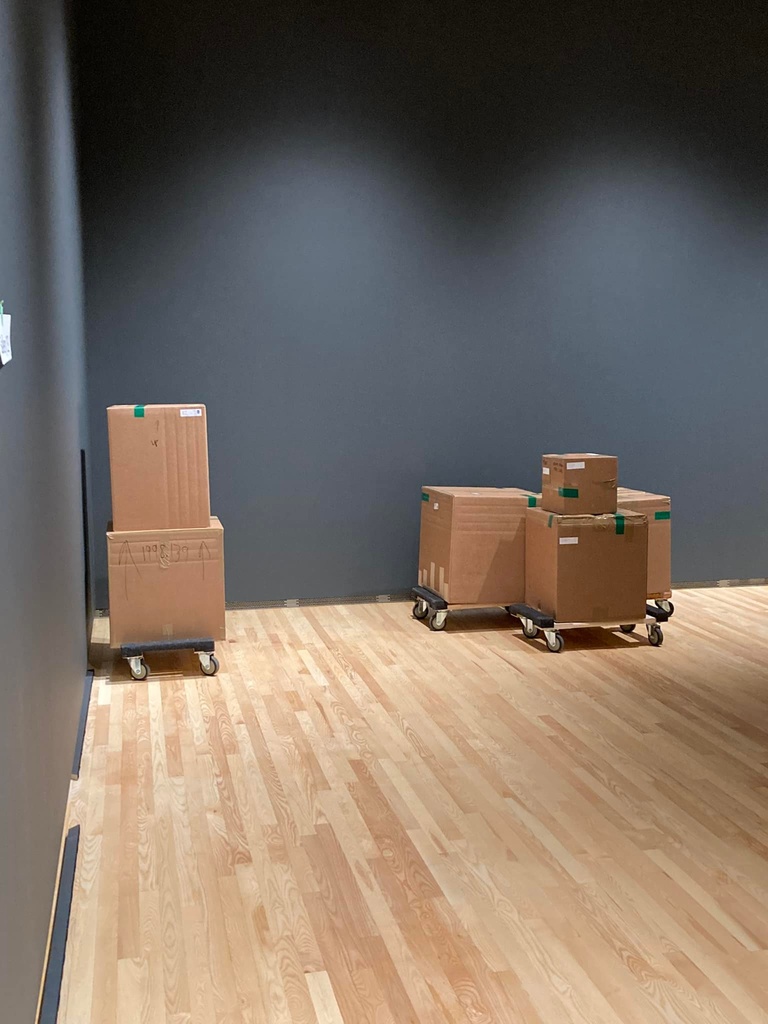 cardboard shipping boxes sit on wheeled carts in an art gallery with dark grey walls.