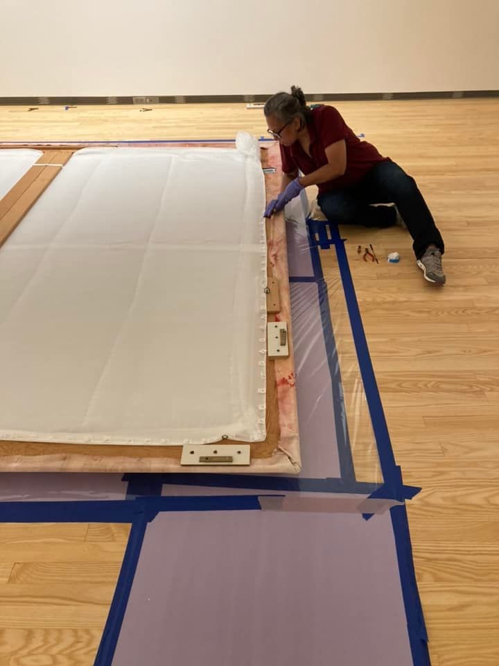 Next to a large painting lying face down on the floor, an art conservator sits and removes staples from the back of the work