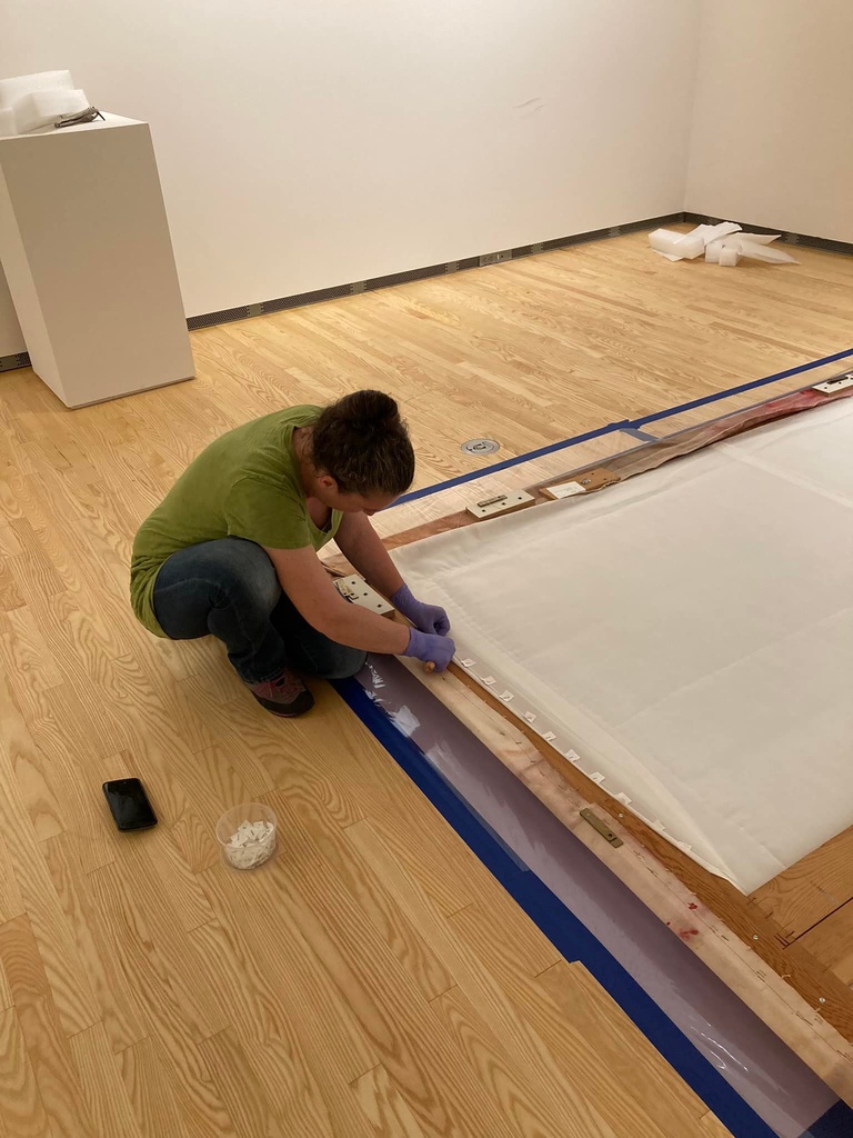 Next to a large painting lying face down on the floor, an art conservator kneels and removes staples from the back of the work