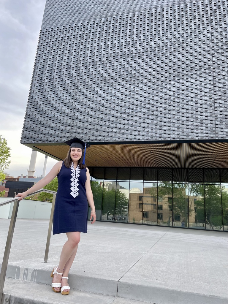 Woman in short navy dress with graduation mortarboard (cap) stands on the front steps of a grey brick building