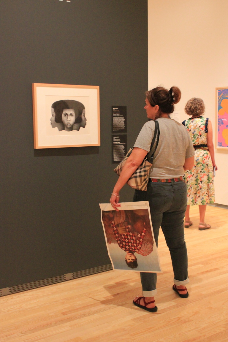 A woman stands looking at a framed lithograph. She holds an art poster in her hand.
