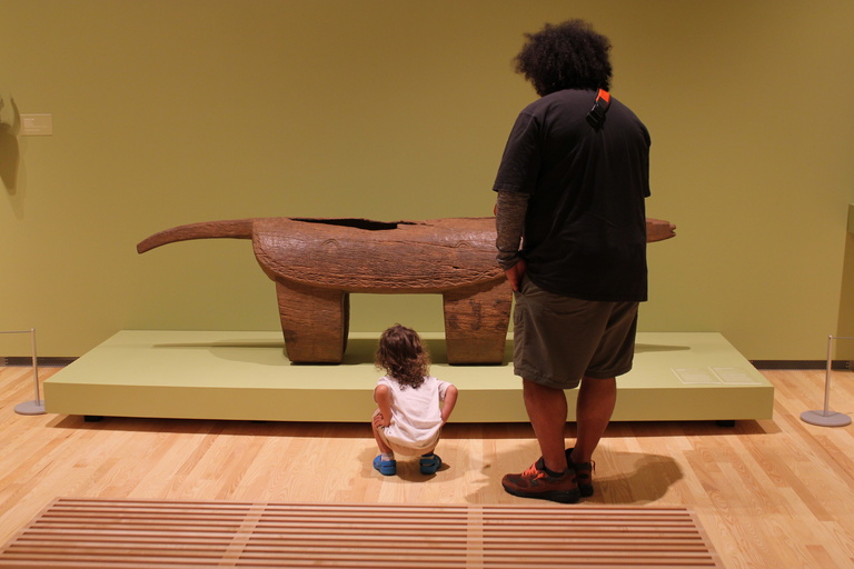 A small child squats in front of a wooden African slit drum. An adult stands next to him.