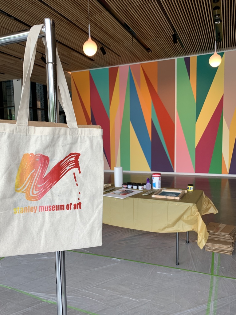 An image of a freshly-screenprinted tote bag hanging on a coat rack. Behind the bag, you can see the table of screenprinting supplies and the Stanley's lobby mural by artist Odili Donald Odita.