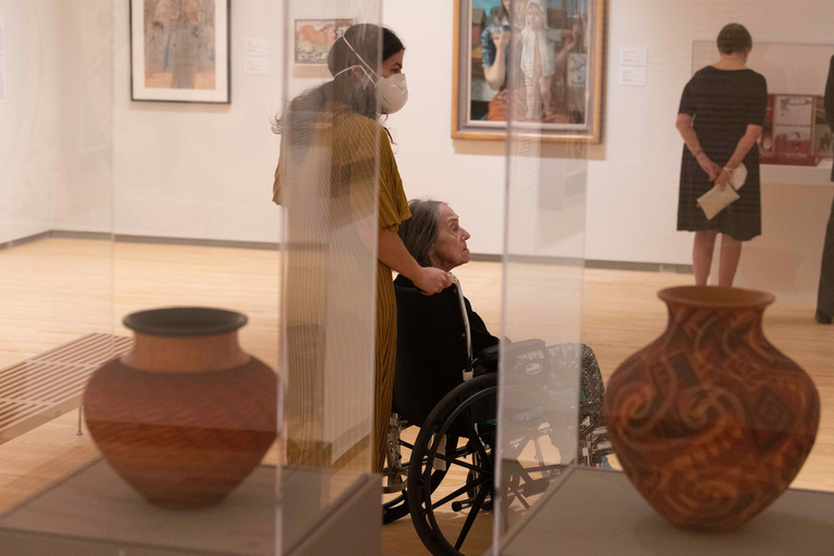 Two people, one in a wheelchair and the other pushing the wheelchair, are seen looking at art in the Stanley's galleries. In the foreground of the shot are two glass cases with clay vessels inside.