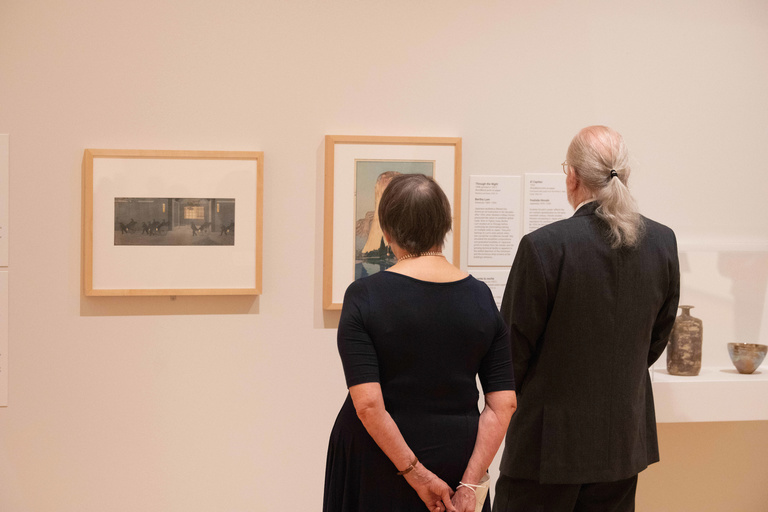 Two people stand, backs to the viewer, looking at framed prints on the wall of the gallery at the Stanley Museum of Art