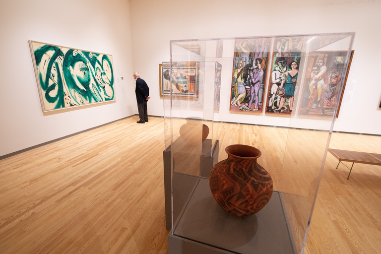 A wide angled shot of a corner of the Stanley Museum of Art gallery. In the corner of the room, a person stands, examining the label next to a large, abstract, green and white painting by Lee Krasner, on loan for the inaugural exhibition.