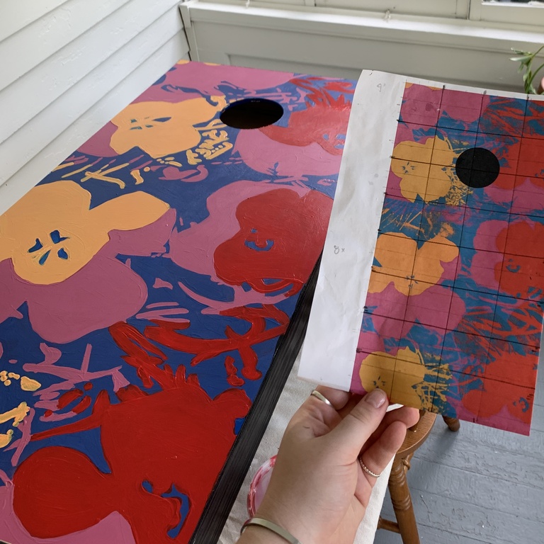 A photo of the painting process for one of the cornhole boards. A hand holds a marked-up, printed image of Andy Warhol's "Flowers" above a cornhole board on which the same design has been painted.