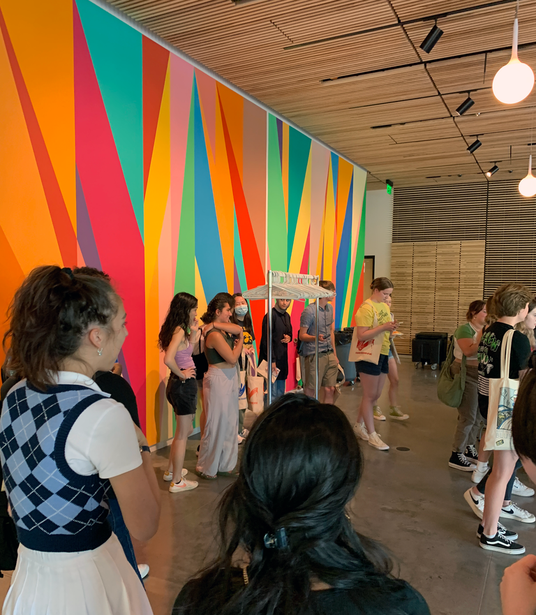 Students wait in line near the Odili Donald Odita mural in the Stanley Museum of Art lobby. They stand near an empty coat rack, with around one hundred hangers on it that once held tote bags.