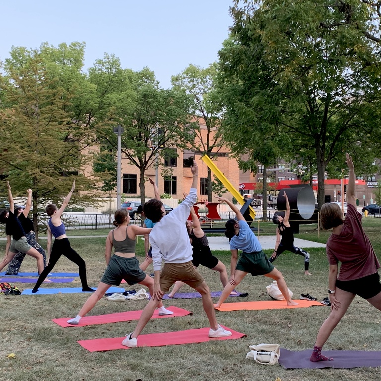 Students doing yoga in Gibson Square Park. The photo is taken from the side, facing Madison Street. There is a hint of outdoor sculpture visible behind the students.