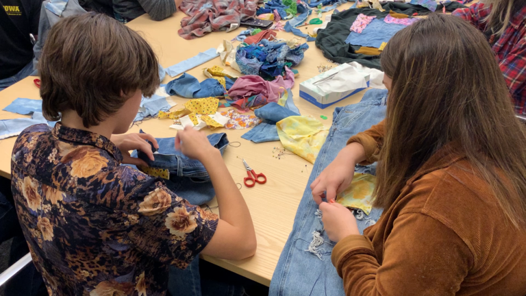 An image of two students sitting together, both bent over their work. The one on the left pulls thread through a garment that they are patching. The one on the right adjusts and makes stitches into the garment she is mending; a pair of ripped jeans with a yellow floral patch she's added.