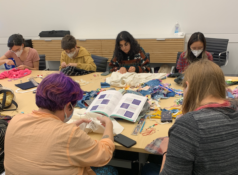 Students are gathered around a conference table, each working on their own mending project. Two students in the foreground, backs to the camera, are focused on a book that is open, displaying various sashiko stitching techniques and patterns.