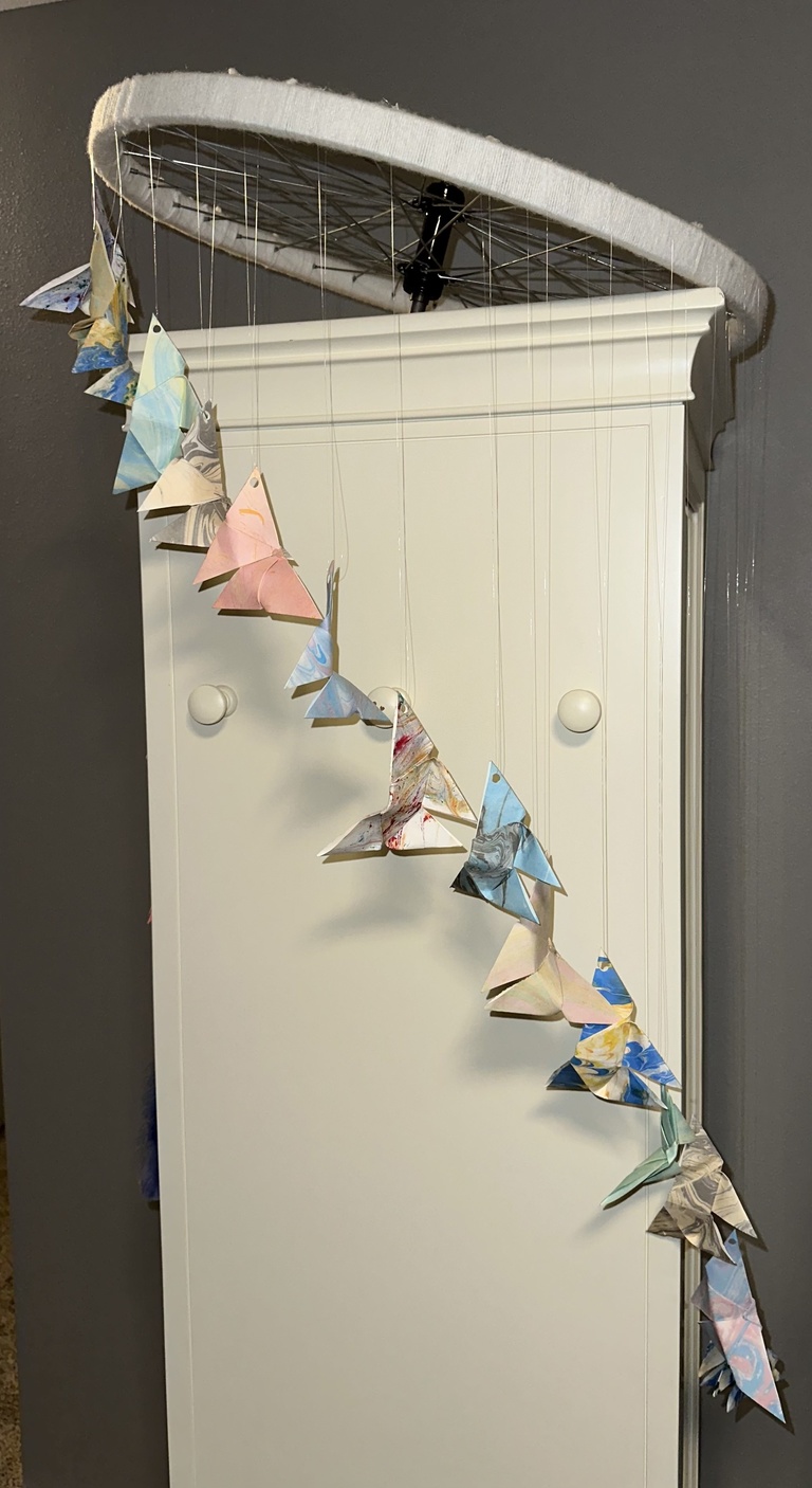 A photo of Jenna Mosnik's art: a mobile suspended from a bicycle wheel, with origami cranes. 