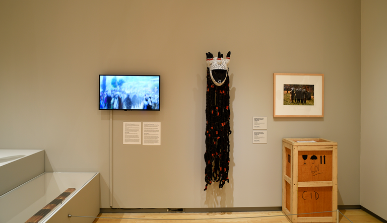 An installation shot of Herve Youmbi's mask, crate, video, photograph, and extended label in "About Face"