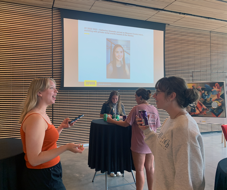 A photo taken at the HBD, SMA event. Two students in the foreground are having a conversation; behind them, other students are at a cocktail table eating cupcakes. In the background is a projector screen with a powerpoint displaying images of student events.