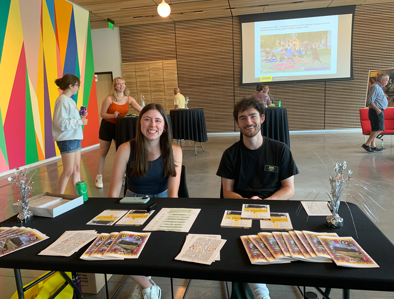A photo of Anaka and Jack at the HBD, SMA information desk. They are smiling, preparing to hand out zines and Student Challenge cards to event visitors.