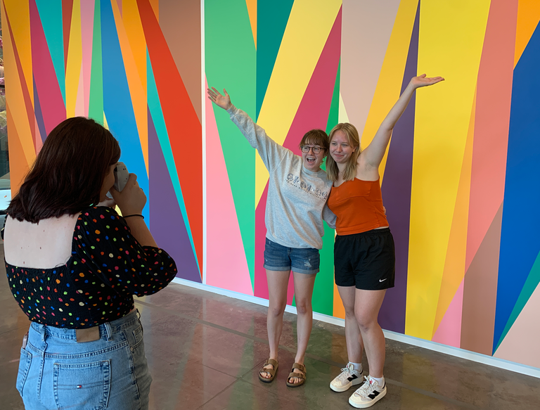 One student takes a Polaroid picture of two other students, posing excitedly, their arms raised, in front of the Stanley's rainbow-colored lobby mural.