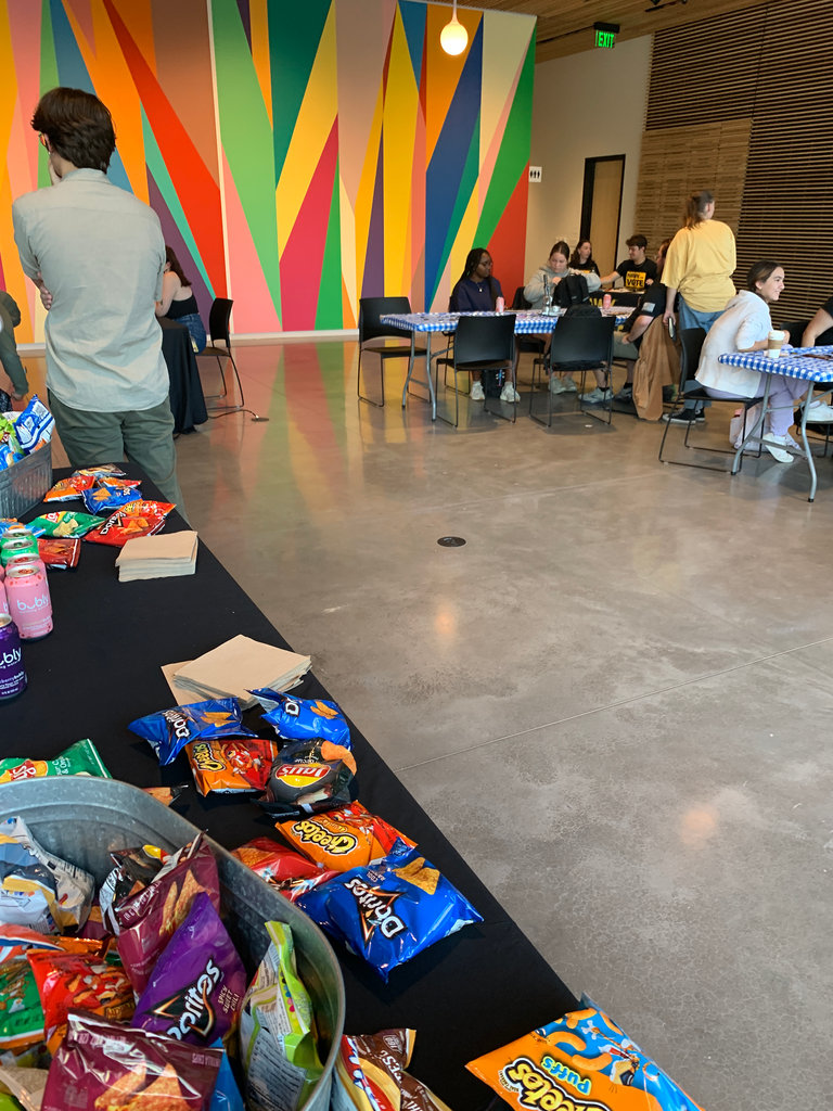 A photo of the event showing a table of snacks, including various bags of chips and sparkling water, with students in the background sitting at tables.