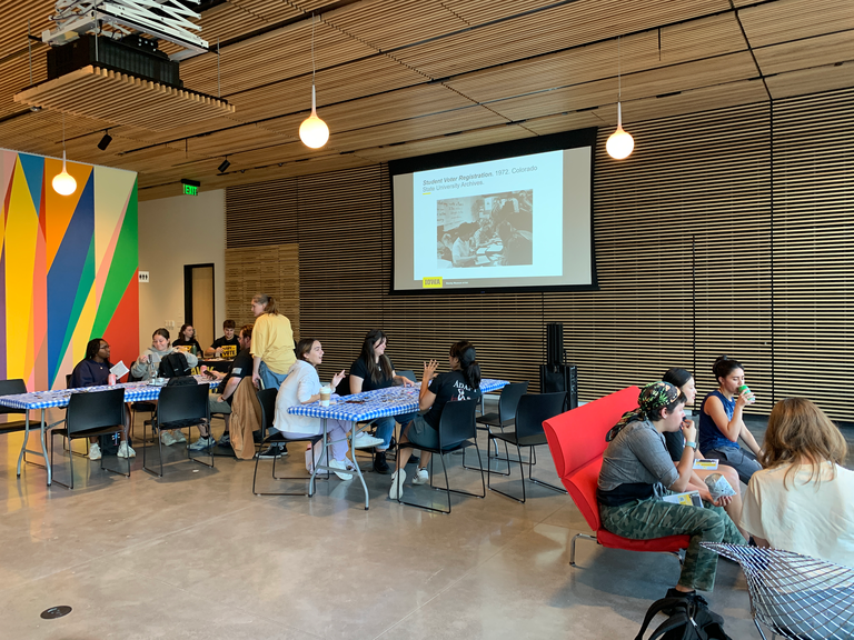 A photo showing the setup of the Art of Voting event--the projector at the far end of the lobby has a looping PowerPoint with images of art and archival photos; there are two tables for button making; and Hawk the Vote has a table to register people to vote.