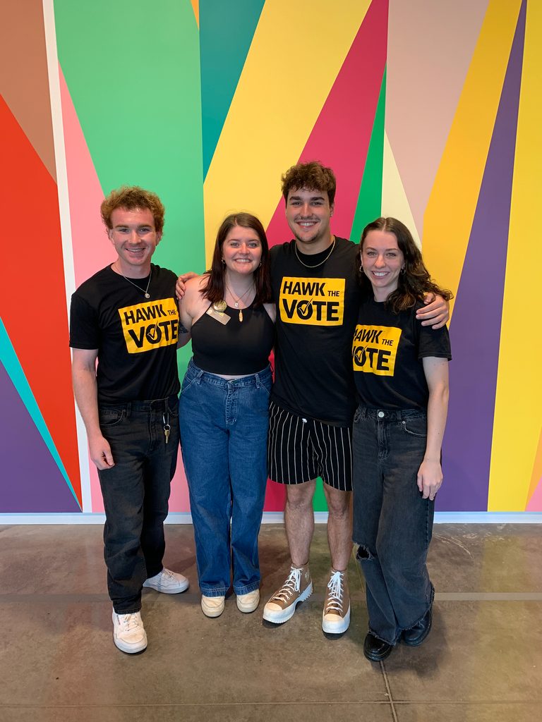 A photo of four students posing together in front of the mural in the Stanley lobby--three students are members of Hawk the Vote, and one is the Stanley's Campus Engagement Coordinator, Alexis.