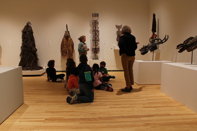 a group of children sit on the floor in front of African masks in a museum gallery