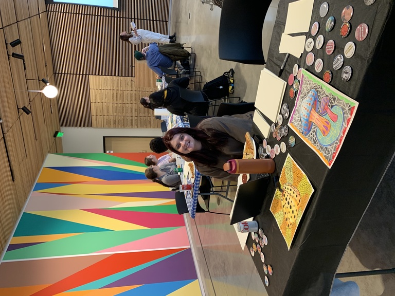picture of campus engagement coordinator Alexis Belme sitting at the welcome table for the event. In the background are students listening to Amanda speaking about the activity 