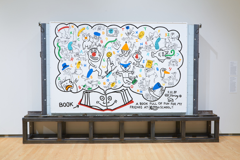 A large white mural filled with colorful drawings, mounted on a concrete wall held together by a metal base
